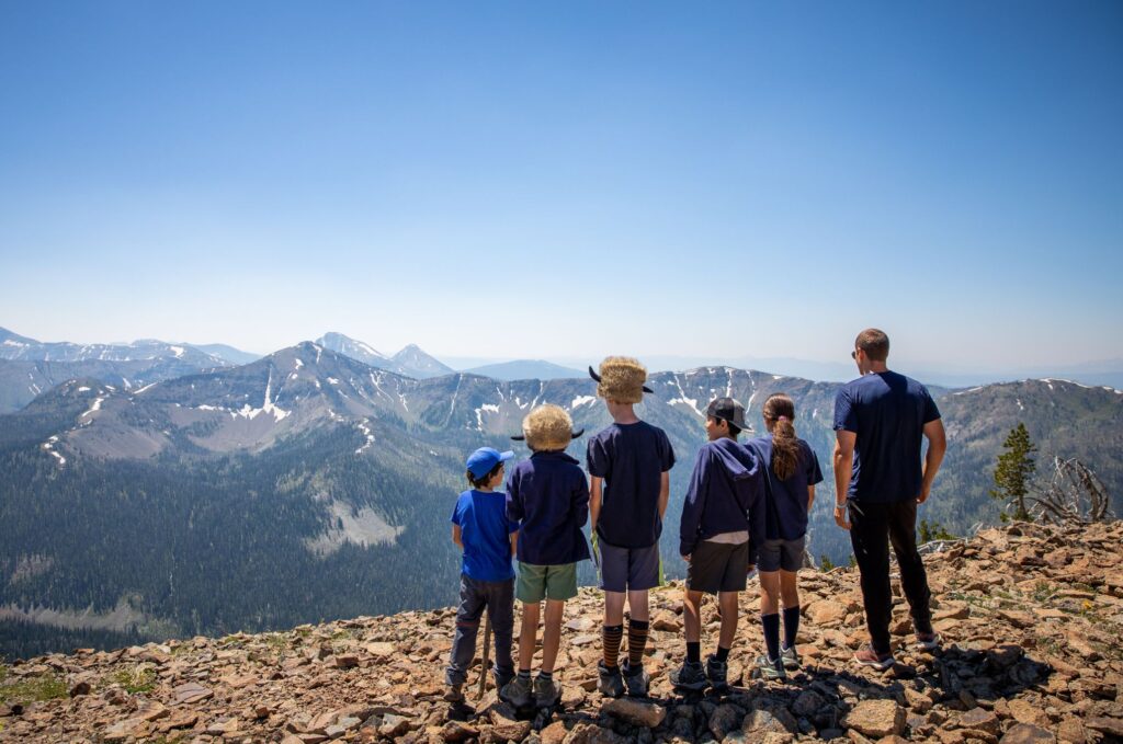 Group of kids and a guide stand on a mountain summit, admiring the panoramic view of rugged alpine peaks.