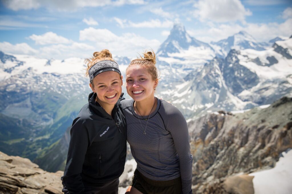 Two friends smile at a snowy mountain viewpoint, with rugged alpine peaks and the Matterhorn in the background.