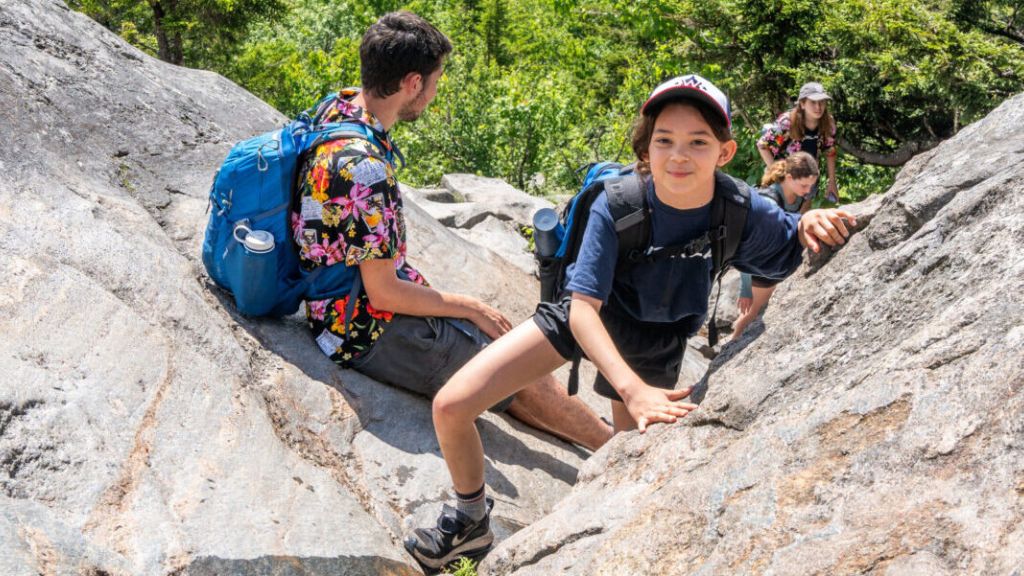 Kids and teens climb rocks in a forest during a summer hike, showcasing teamwork and fun at Adventure Camps in the great outdoors.