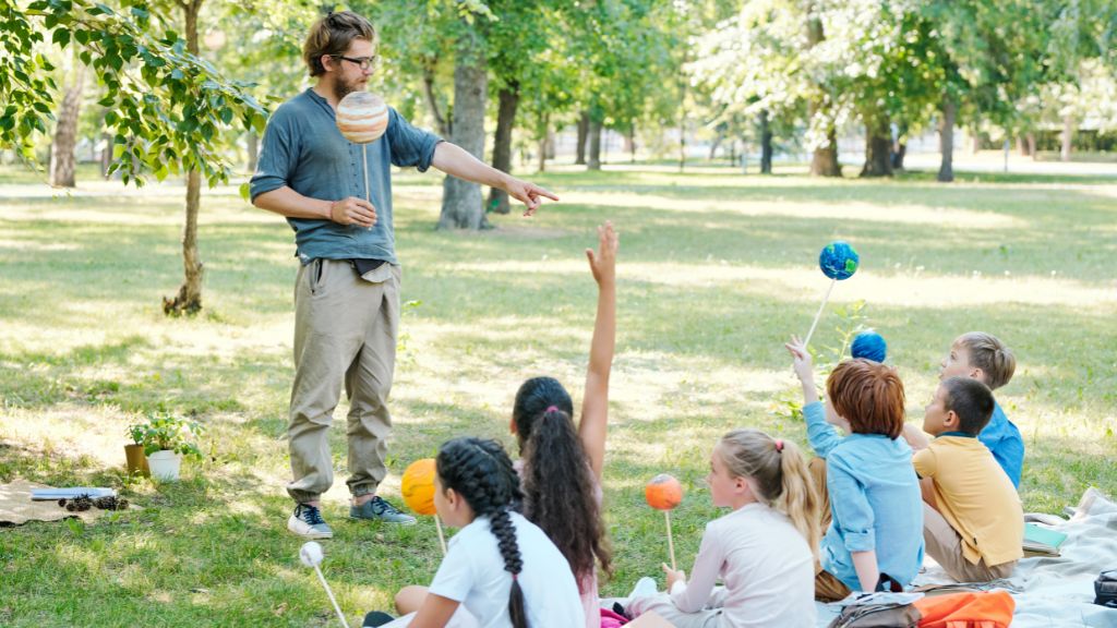 A group of curious kids sits on the grass holding planet models as a teacher leads a fun science lesson at Day Camps in a sunny park setting.
