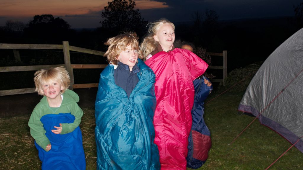 Smiling kids bundled in sleeping bags enjoy a fun evening outdoors at Traditional Overnight Camps, creating memories under the stars.