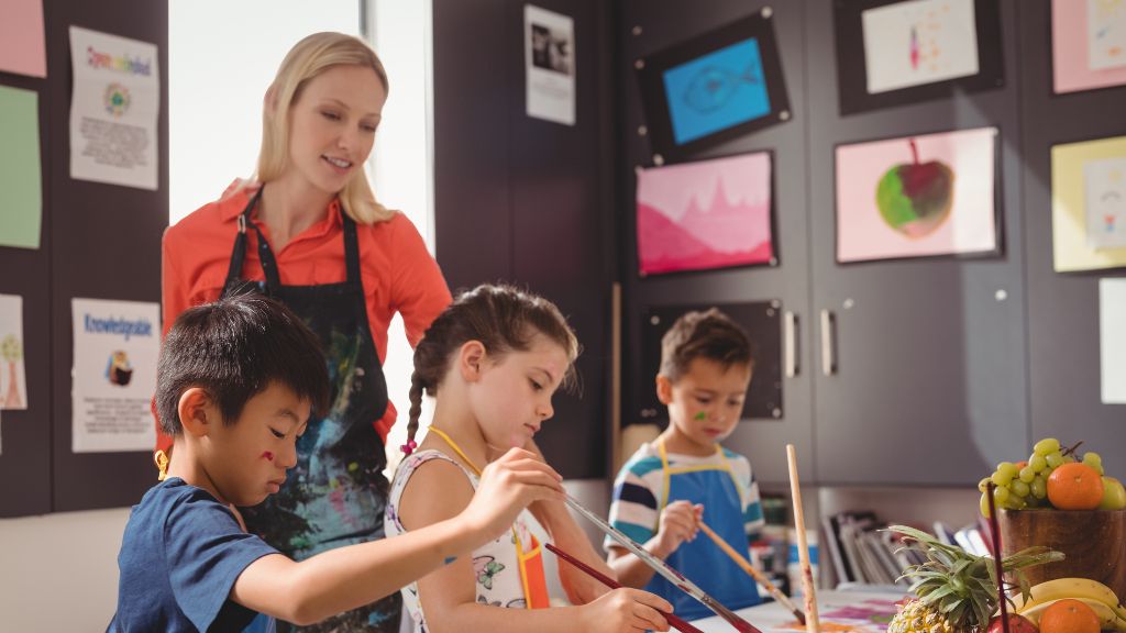 Children paint with focus and creativity in a colorful classroom as a teacher offers guidance, capturing the spirit of expression at Arts Camps.
