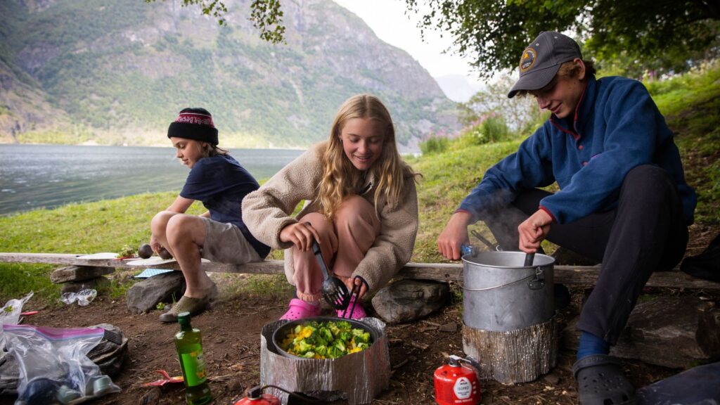 Campers cooking outdoors beside a lake with guidance from staff, learning leadership and life skills at summer camp.