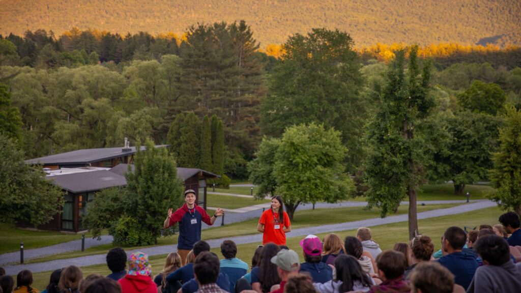 Camp counselors leading an orientation talk with campers seated outdoors at summer camp, surrounded by cabins and trees at sunset.