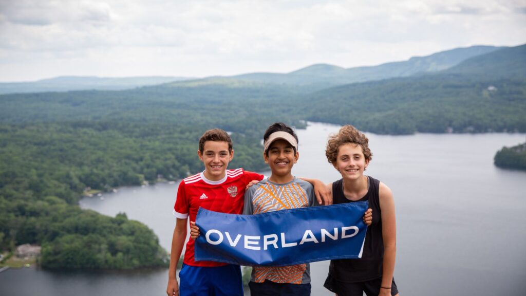 Three smiling teens holding an Overland banner atop a scenic overlook, representing Summer Travel Programs for Teens in nature