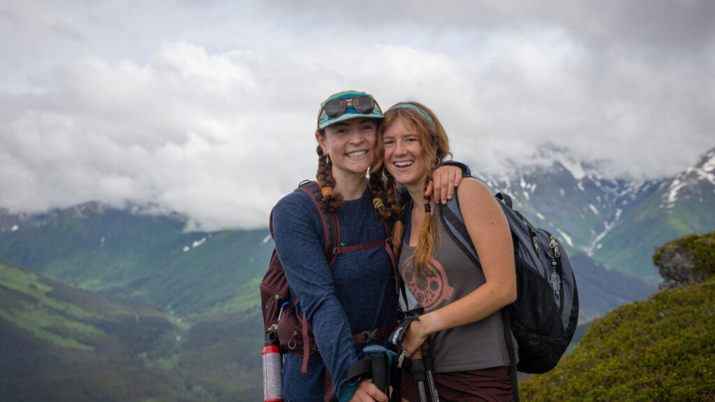 Two smiling teens with backpacks standing on a mountain trail with cloudy peaks and green valleys behind them during the Alaska Expedition Summer Travel Program for Teens.