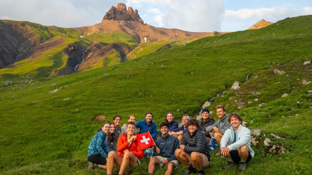 Group of teens sitting on a grassy alpine slope with a rocky peak in the background, holding a Swiss flag during the Alpine Challenge Summer Travel Program for Teens.