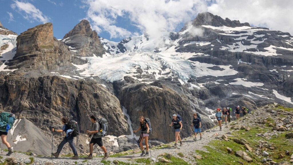 Group of teens hiking along a mountain trail with glaciers and rugged peaks in the background during the Alpine Explorer Summer Travel Program for Teens.