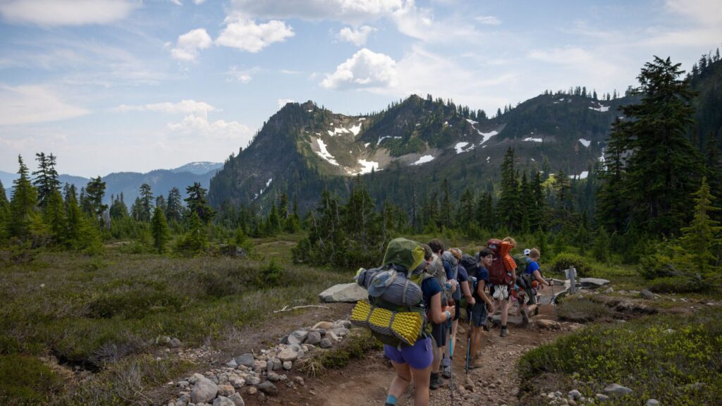 Teens backpacking along a mountain trail with snow-capped peaks in the distance, part of the Northwest Explorer Summer Travel Program for Teens.