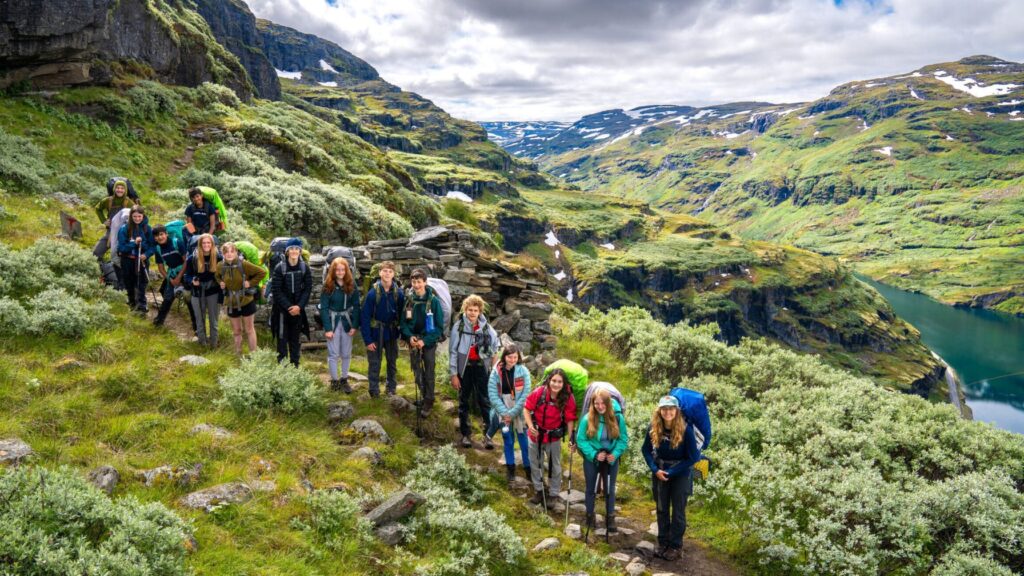 Group of teens with backpacks hiking through lush green mountains and valleys with lakes below during the Norway Expedition Summer Travel Program for Teens.