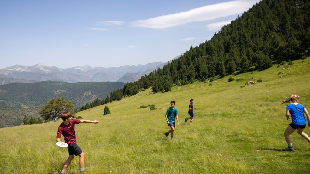 Teens playing frisbee in a grassy meadow surrounded by forested hills and mountains during the Pyrenees Expedition Summer Travel Program for Teens.