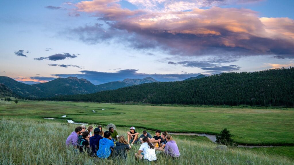 Group of teens sitting in a grassy field with mountains in the background at sunset, participating in the Rocky Mountain Explorer Summer Travel Program for Teens.
