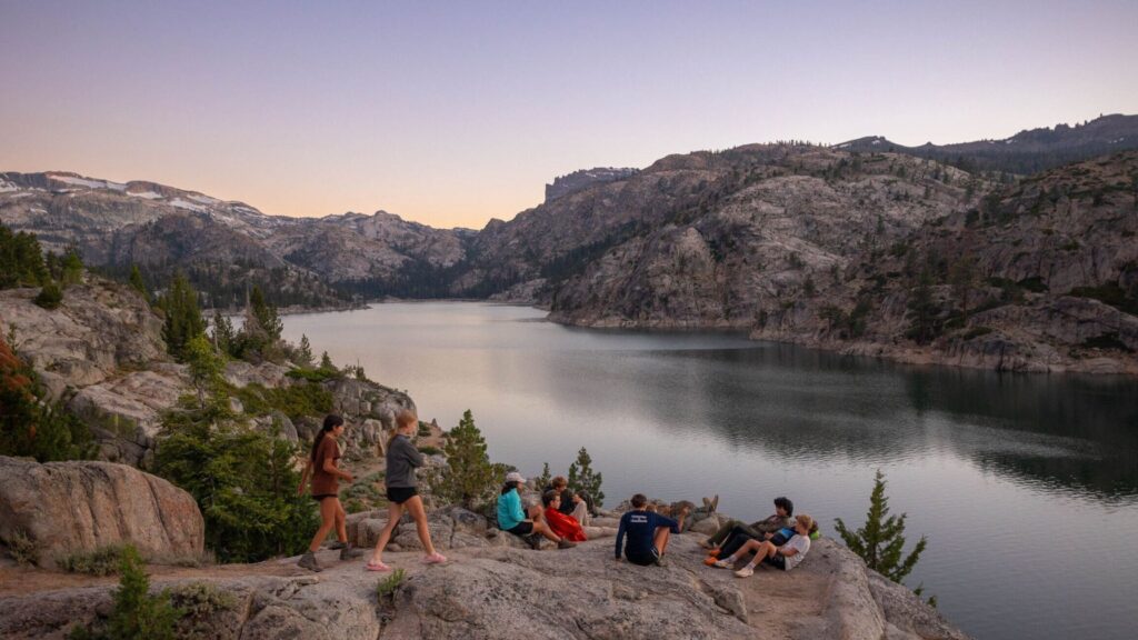 Teens relaxing on rocky cliffs overlooking a lake surrounded by granite mountains during the Sierra Explorer Summer Travel Program for Teens.
