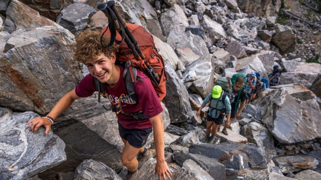 Teens climbing over rocky terrain with backpacks and hiking poles during the Teton Challenge Summer Travel Program for Teens.