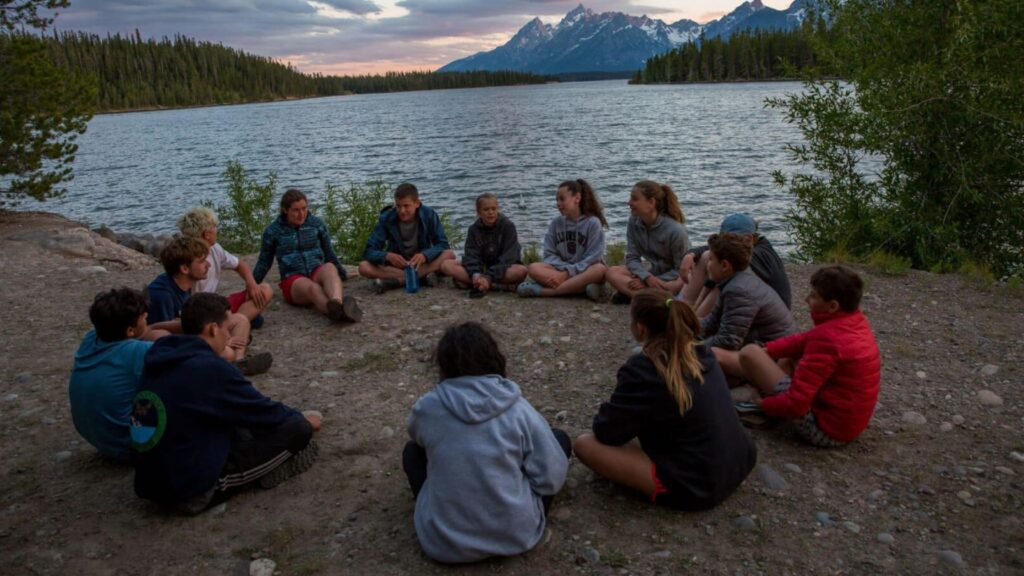 Teens sitting in a circle by a lake at sunset with mountains in the background during the Yellowstone Teton Explorer Summer Travel Program for Teens.