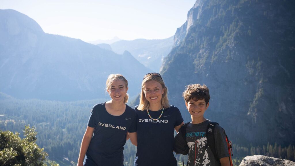 Three kids ages 10 to 12 smile at the camera during a summer camp hike, standing in front of a scenic mountain landscape, wearing Overland shirts and backpacks.