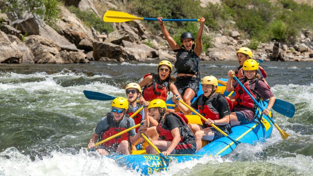 Teens ages 13 to 15 cheer and paddle together while whitewater rafting through rapids during a summer camp leadership program, wearing helmets and life vests.