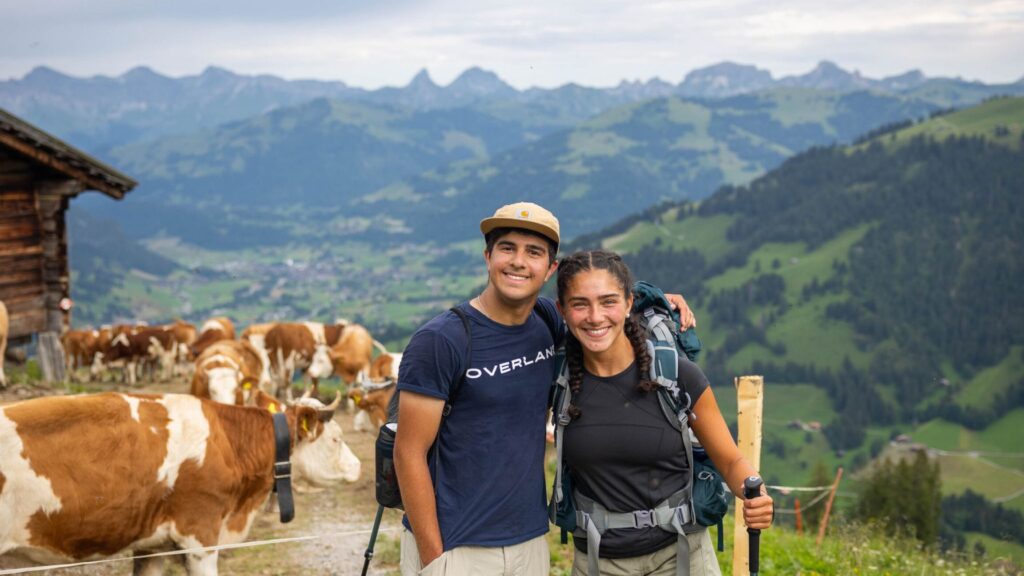 Two teens ages 16 and up smile while hiking through the mountains during a pre-college summer camp experience, with backpacks on and cows grazing in the background.