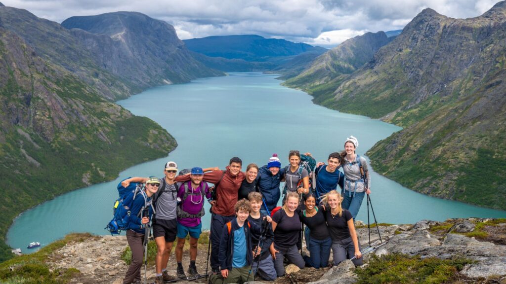 A group of teenage campers pose together at the edge of a scenic cliff overlooking a turquoise lake and surrounding mountains during a summer camp hiking expedition.
