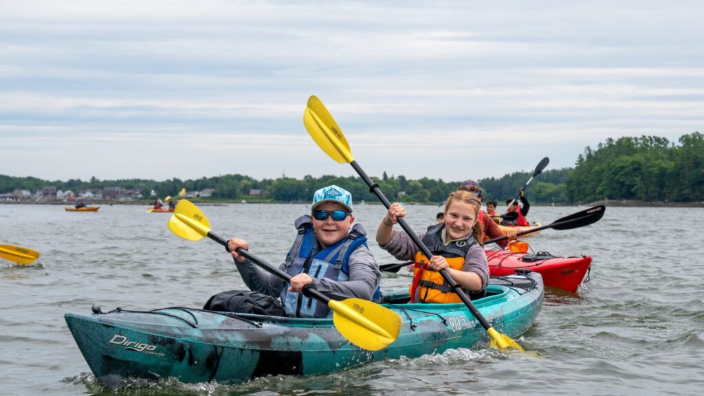 Two kids kayaking on a calm lake during a screen-free summer, smiling and paddling together with a group in the background under cloudy skies.