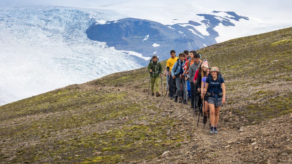 A group of hikers walking in a line along a rocky mountain trail with trekking poles, with a vast glacier and rugged peaks in the background.