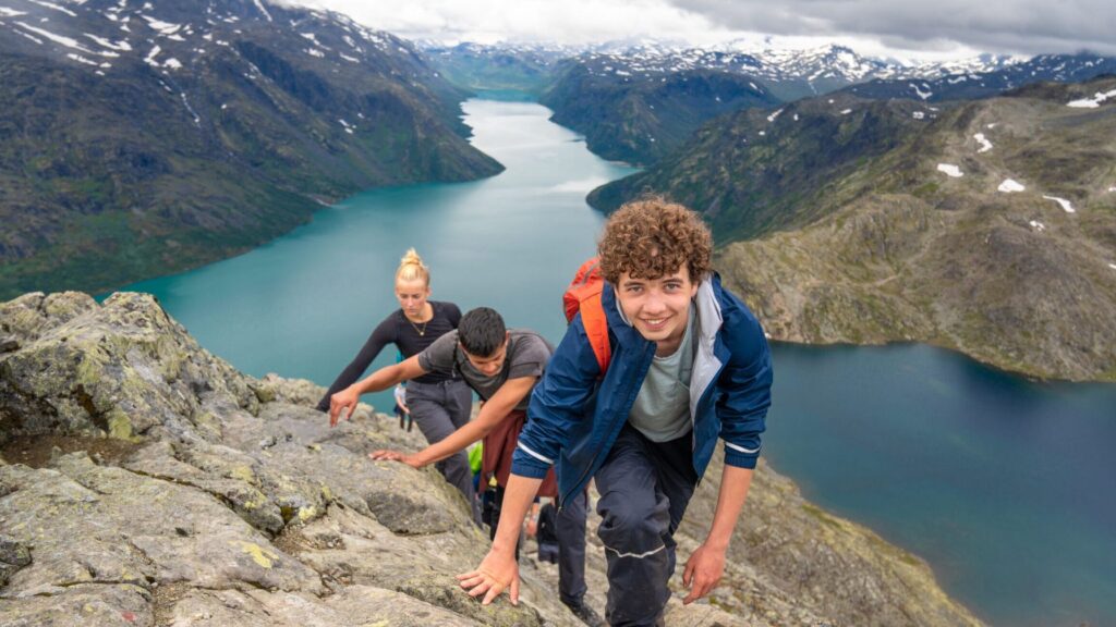 Teens climbing a rocky mountain ridge during a screen free summer adventure, with a stunning lake and mountain valley in the background.