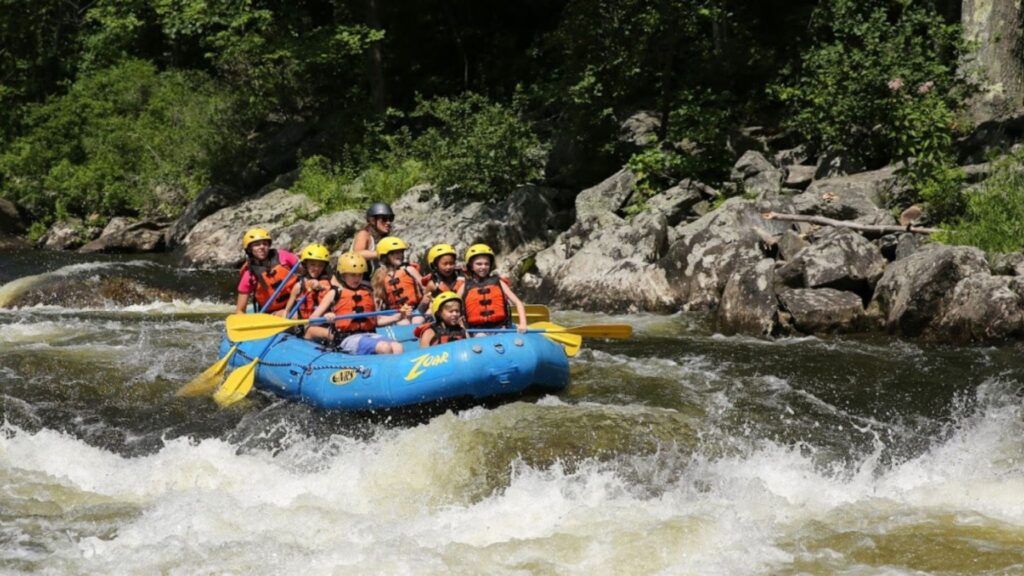 Children ages 6 to 9 wearing life jackets and helmets whitewater rafting in a blue inflatable raft during a summer sleepaway camp adventure.