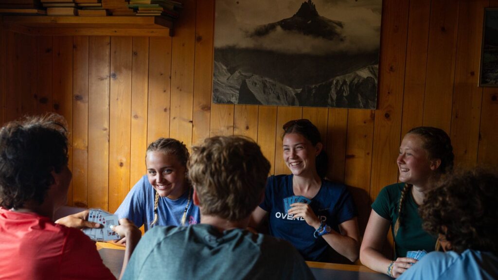 A group of smiling campers sit around a wooden table inside a cozy cabin, playing cards and sharing laughs during downtime at summer camp.