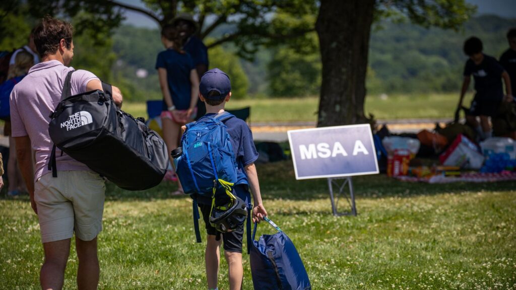 A father and son carrying bags arrive at a summer camp check-in area marked 'MSA A,' with other campers and supplies visible under a tree in the background.