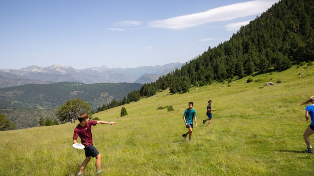 Campers play a game of frisbee in a wide open grassy field surrounded by forested hills and distant mountain ranges, highlighting a scenic summer camp environment.