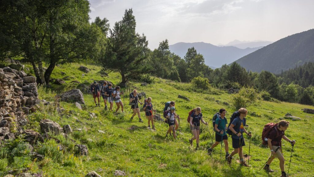 A group of campers hike in a single-file line through a lush green mountain landscape, each carrying backpacks and trekking poles during a summer camp adventure.