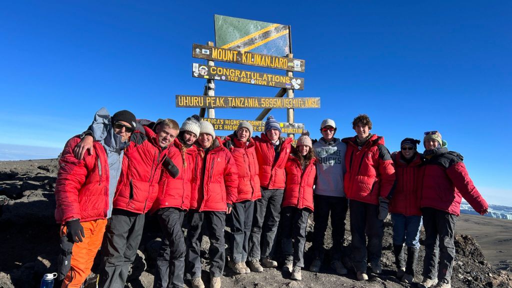 Group in red jackets stand together at a mountain summit sign, celebrating reaching the peak under a clear blue sky.