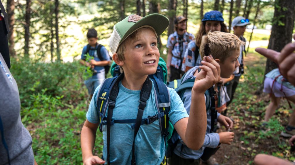 Kids hike through a forest trail; one boy in front smiles and holds up a small object while others gather nearby.