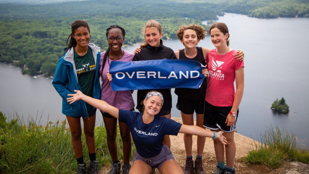 Overland campers and trip leader posing with a banner at a scenic lake overlook.